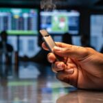 Adult hand holding a slim pod-style vape with no lights and minimal vapor above a bar counter, with softly blurred sportsbook TV screens and patron silhouettes in the background.